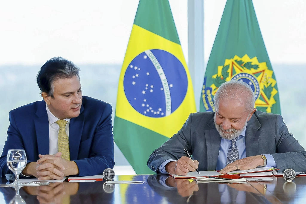 Na imagem, dois homens estão sentados à mesa em um ambiente formal. Um deles, à direita, está assinando um documento, enquanto o outro, à esquerda, observa. Ao fundo, estão as bandeiras do Brasil, uma com o brasão e a outra com o símbolo nacional. A mesa tem copos de água e canetas visíveis. Presidente Lula (PT) durante reunião com ministro da Educação, Camilo Santana, para assinatura do Decreto
