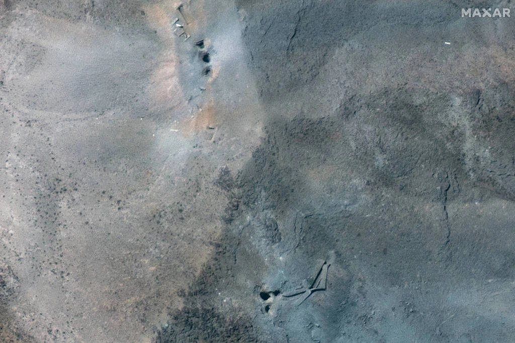 A imagem mostra uma área de superfície terrestre com tons de cinza e marrom, apresentando uma textura irregular. Há algumas marcas ou depressões visíveis no solo, sugerindo atividade ou alterações na superfície. O céu parece claro, e a imagem é capturada em uma perspectiva aérea. Imagem de satélite mostra seis pontos de entrada de bombas em Fordow