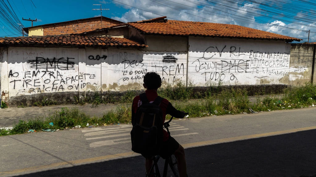 Pessoa com mochila sentada em bicicleta na sombra observa muro branco com várias pichações em preto. O muro tem telhado de cerâmica e está em área urbana com céu parcialmente nublado ao fundo.  Pichações mostram ordens e siglas de facção criminosa em rua de Maranguape, na região metropolitana de Fortaleza (CE); município teve a maior taxa de homicídios estimados no país, com 87,2 casos para cada 1